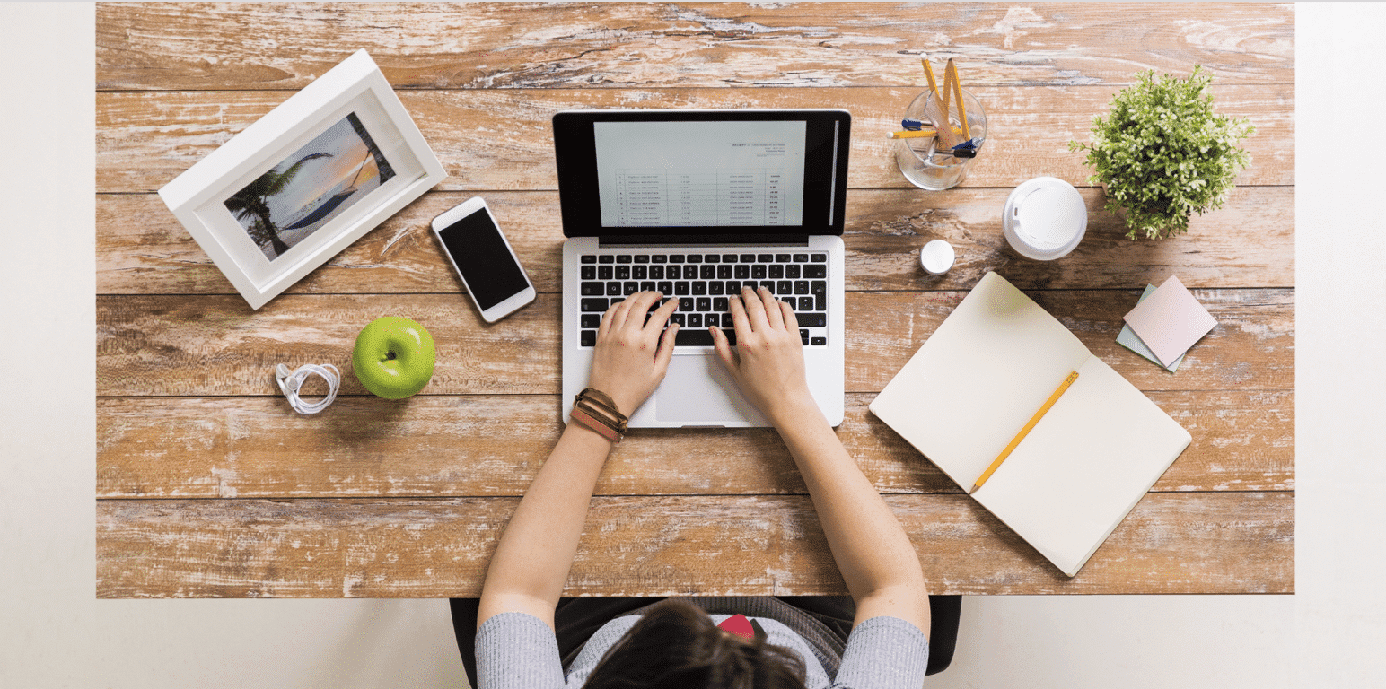 Person typing on laptop at wooden desk with green apple, smartphone, notepad, and decorative items, illustrating a productive workspace for digital marketing activities.