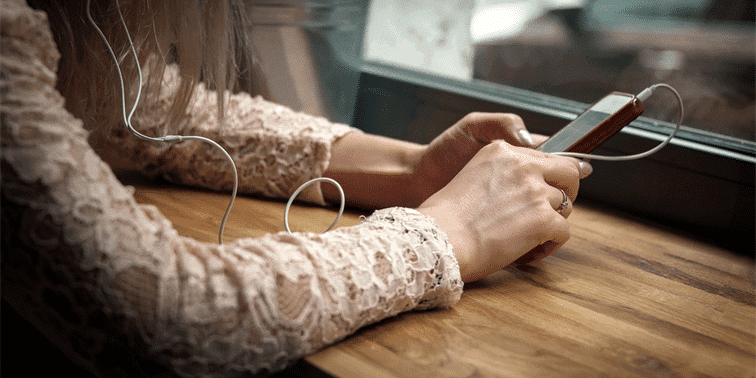Woman using smartphone with earphones, sitting at wooden table, engaged in digital content, relevant to TikTok and video marketing.