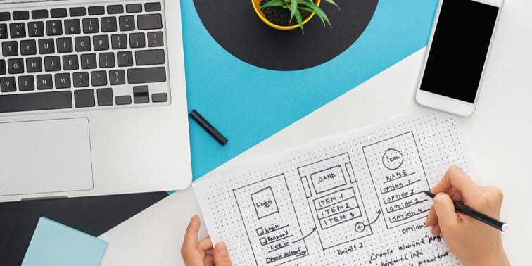 Woman sketching website design wireframes on paper with laptop and smartphone on desk, illustrating web design concepts relevant to digital marketing strategies.